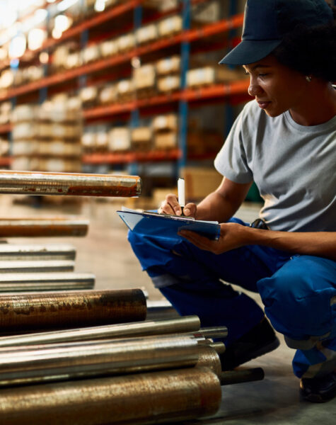 African American female worker taking notes while working in a w Black female worker writing on clipboard while inspecting steel products at industrial warehouse.