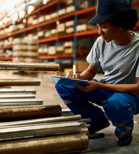 African American female worker taking notes while working in a w Black female worker writing on clipboard while inspecting steel products at industrial warehouse.