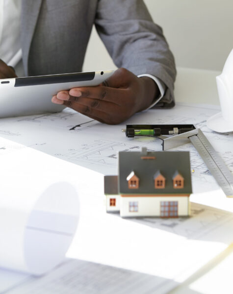 Cropped shot of dark-skinned contractor holding touch pad, entering data while working on new housing project, sitting at desk with drawings, scale model house, blueprint rolls, ruler and helmets Cropped shot of dark-skinned contractor holding touch pad, entering data while working on new housing project, sitting at desk with drawings, scale model house, blueprint rolls, ruler and helmets