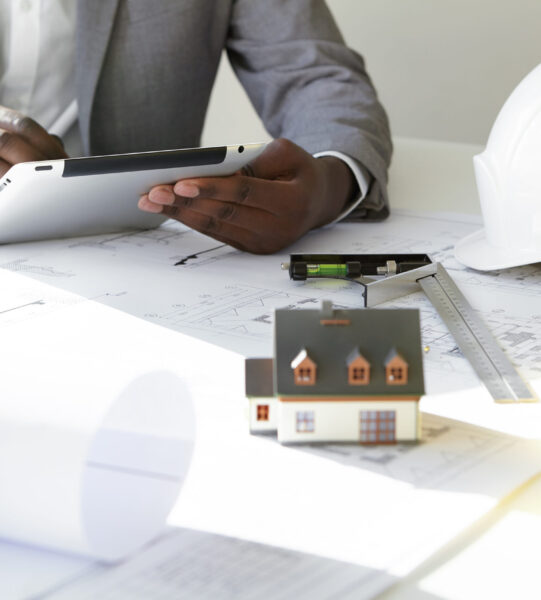 Cropped shot of dark-skinned contractor holding touch pad, entering data while working on new housing project, sitting at desk with drawings, scale model house, blueprint rolls, ruler and helmets Cropped shot of dark-skinned contractor holding touch pad, entering data while working on new housing project, sitting at desk with drawings, scale model house, blueprint rolls, ruler and helmets